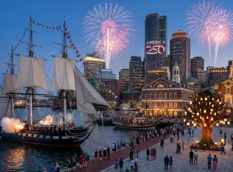 Boston skyline with fireworks and a tall ship with canon fire along the harbor with historic fanuiel hall in the foreground and spectators at water's edge