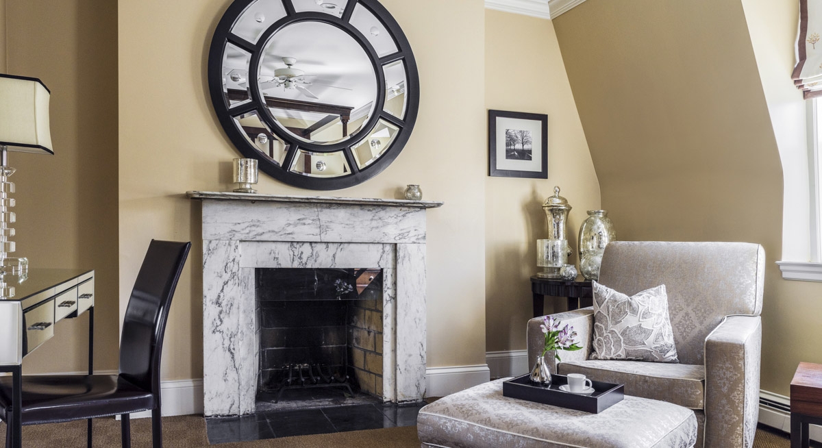 Beige guest room with white marble fireplace, off white upholstered chair and ottoman, with round mirror over mantle.