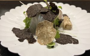 black and white truffles thinly sliced on top of micro green and white fish on a white scallop edge plate