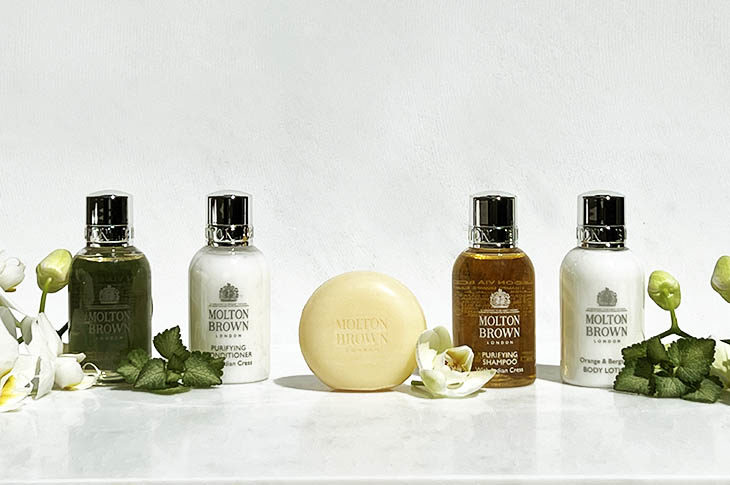 Four bottles of shampoo and a tan bar on soap in the middle with white blossoms and green fern leaves
