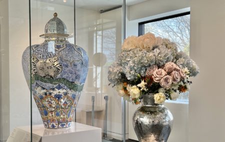 silver vase with purple roses blue hydrogen and white flowers in from of a decorative vase with similar colors in a glass case