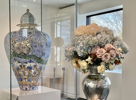 silver vase with purple roses blue hydrogen and white flowers in from of a decorative vase with similar colors in a glass case