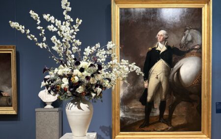 White vase with white flowers next to a gold frame with a painting of George Washing next to a white horse