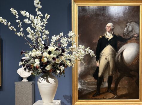 White vase with white flowers next to a gold frame with a painting of George Washing next to a white horse