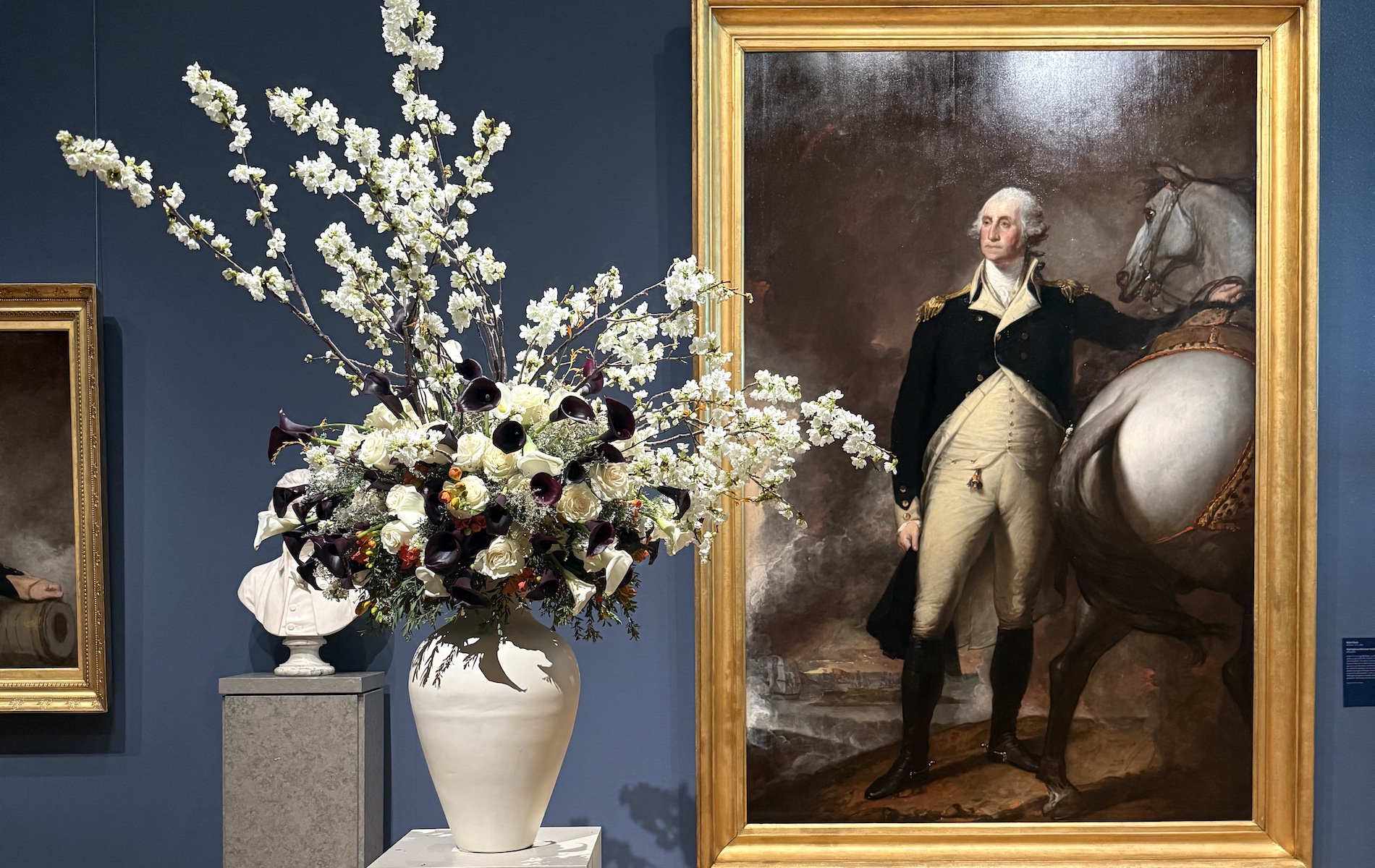 White vase with white flowers next to a gold frame with a painting of George Washing next to a white horse