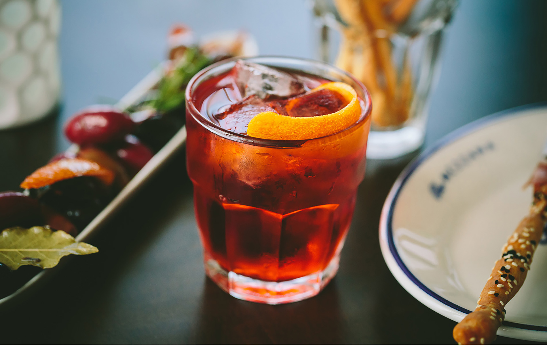 red cocktail with orange slice next to a white plate bread stick