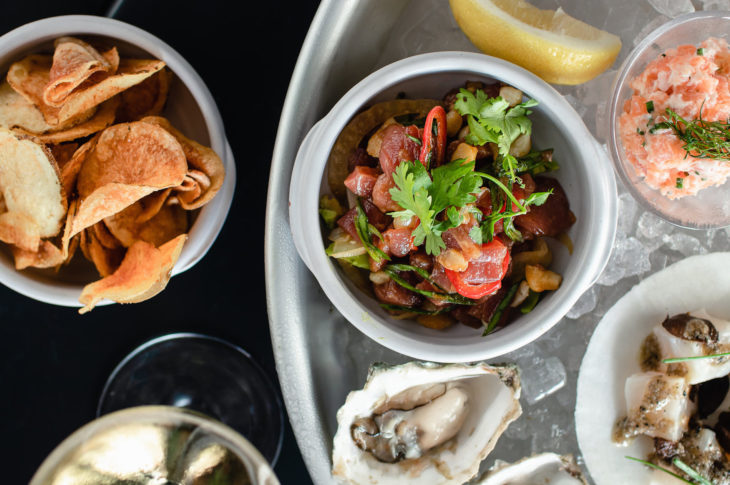 a bowl of orange sweet potato chips next to a tray of ice with 1 oyster 1 lemon wedge and 3 dishes of raw seafood