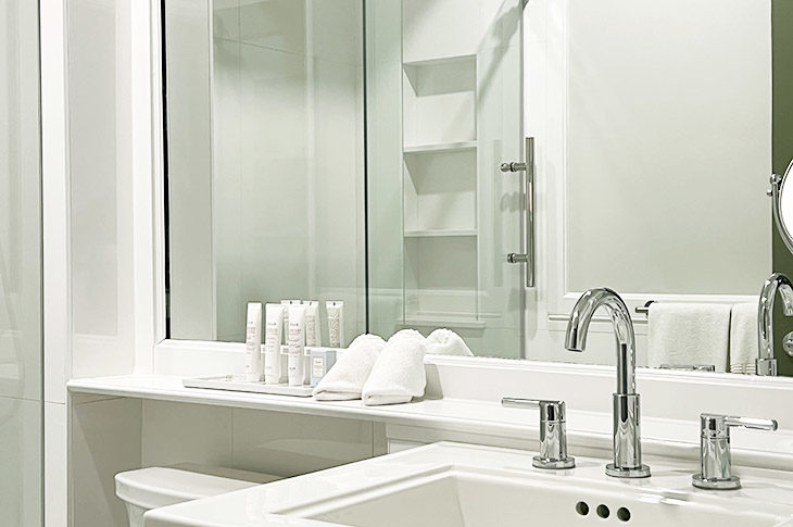 White bathroom with pedestal sink, large mirror and glass tub enclosure.