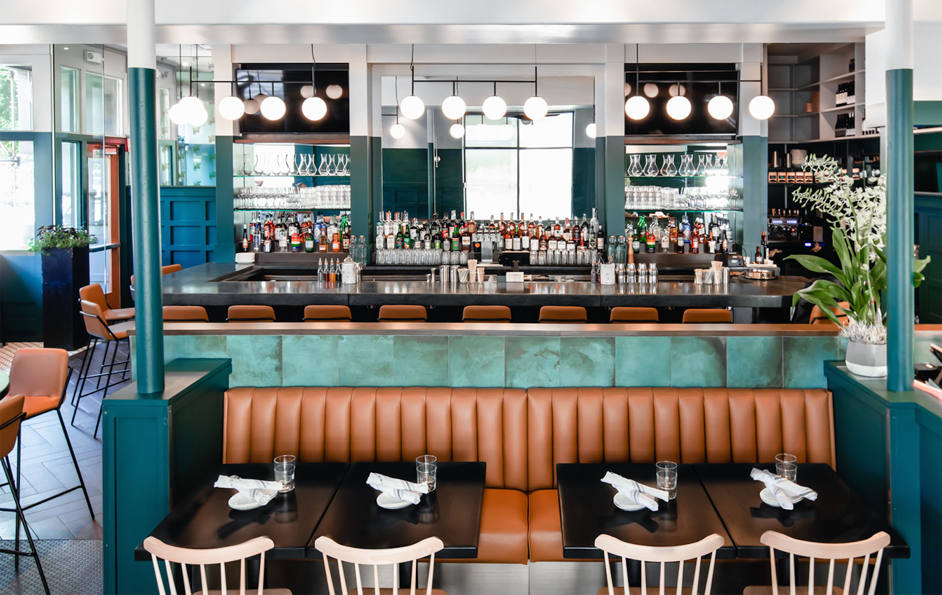 A restaurant with a brown leather booth and earth green walls with a bar behind and a dozen white hanging globe lights