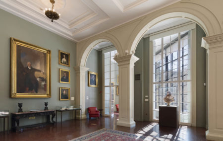 historic room with hight green wall with 5 gold framed artworks and two white arches in front of the whindows