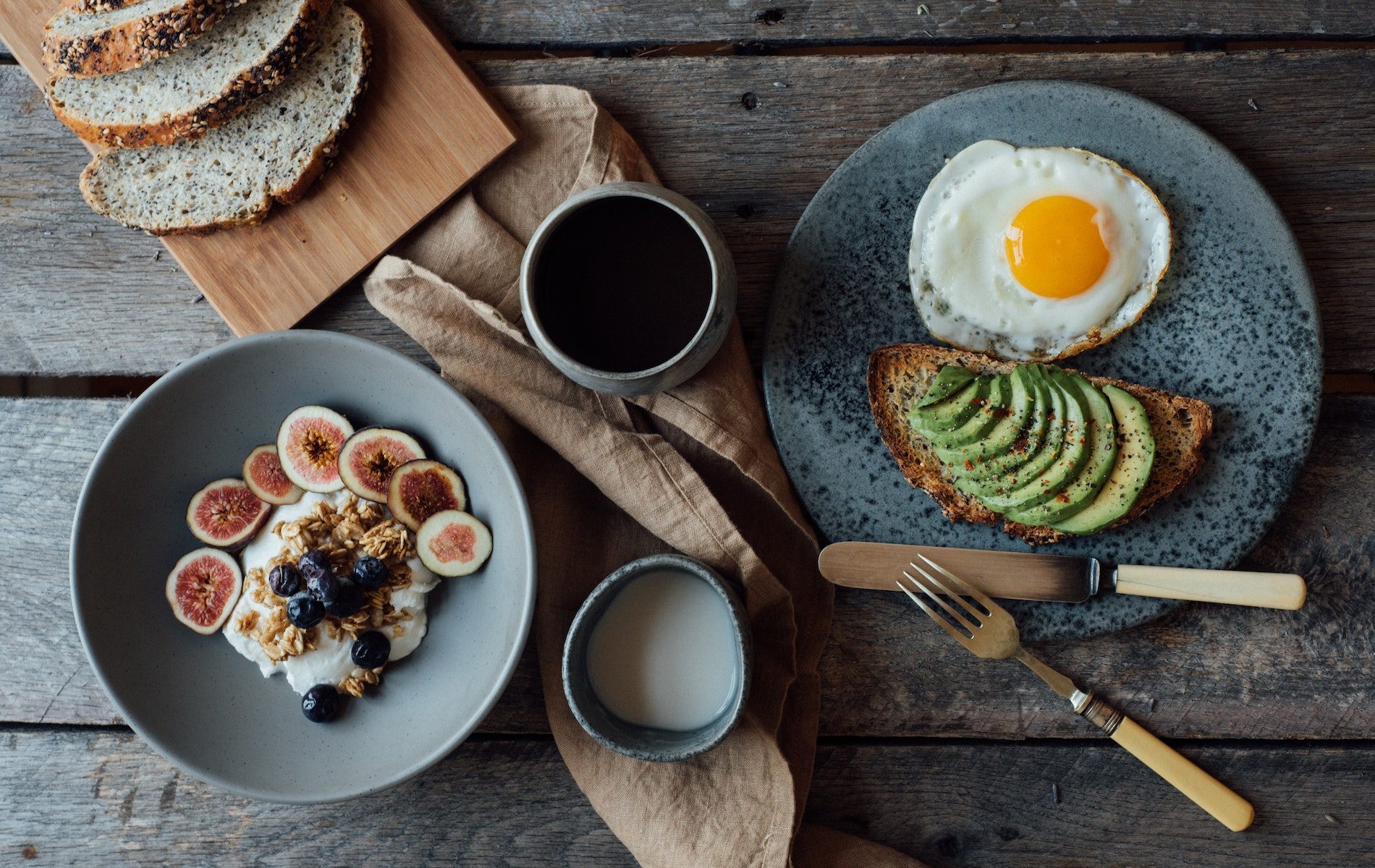 bowl of yogurt with sliced figs brown napkin black coffee cup of milk and avocado slicer with sunny side up egg