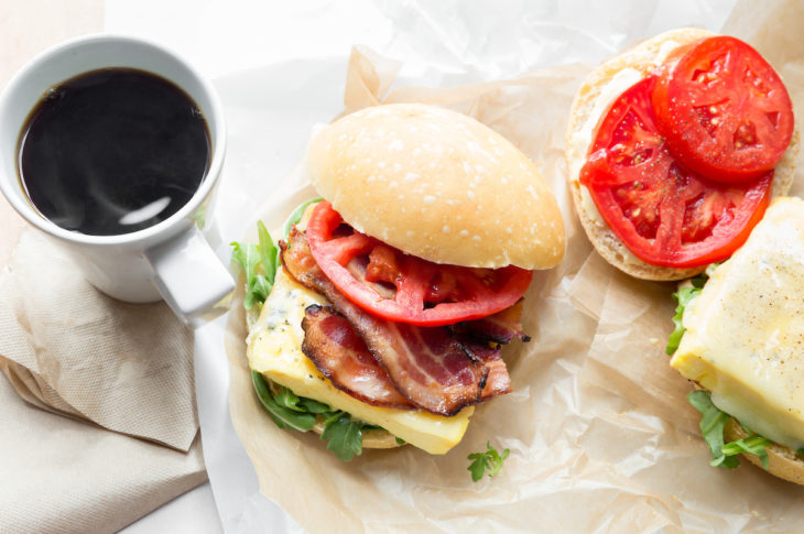 black coffee and a breakfast sandwich with bacon and two slices of red tomato to the right