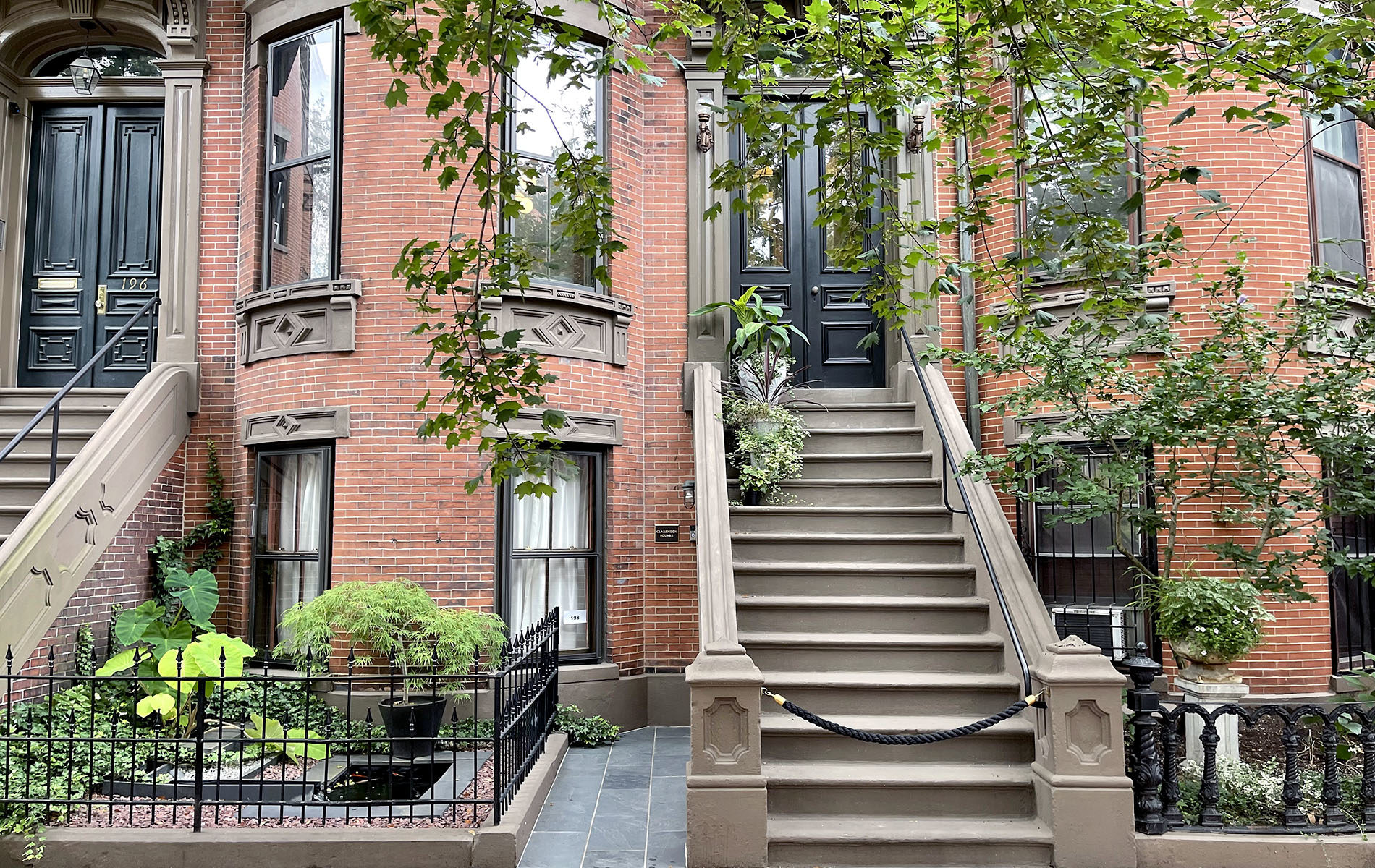 Red brick brownstone with large stone staircase leading to double black entry doors. Tree branches from above and many plants in gardens.