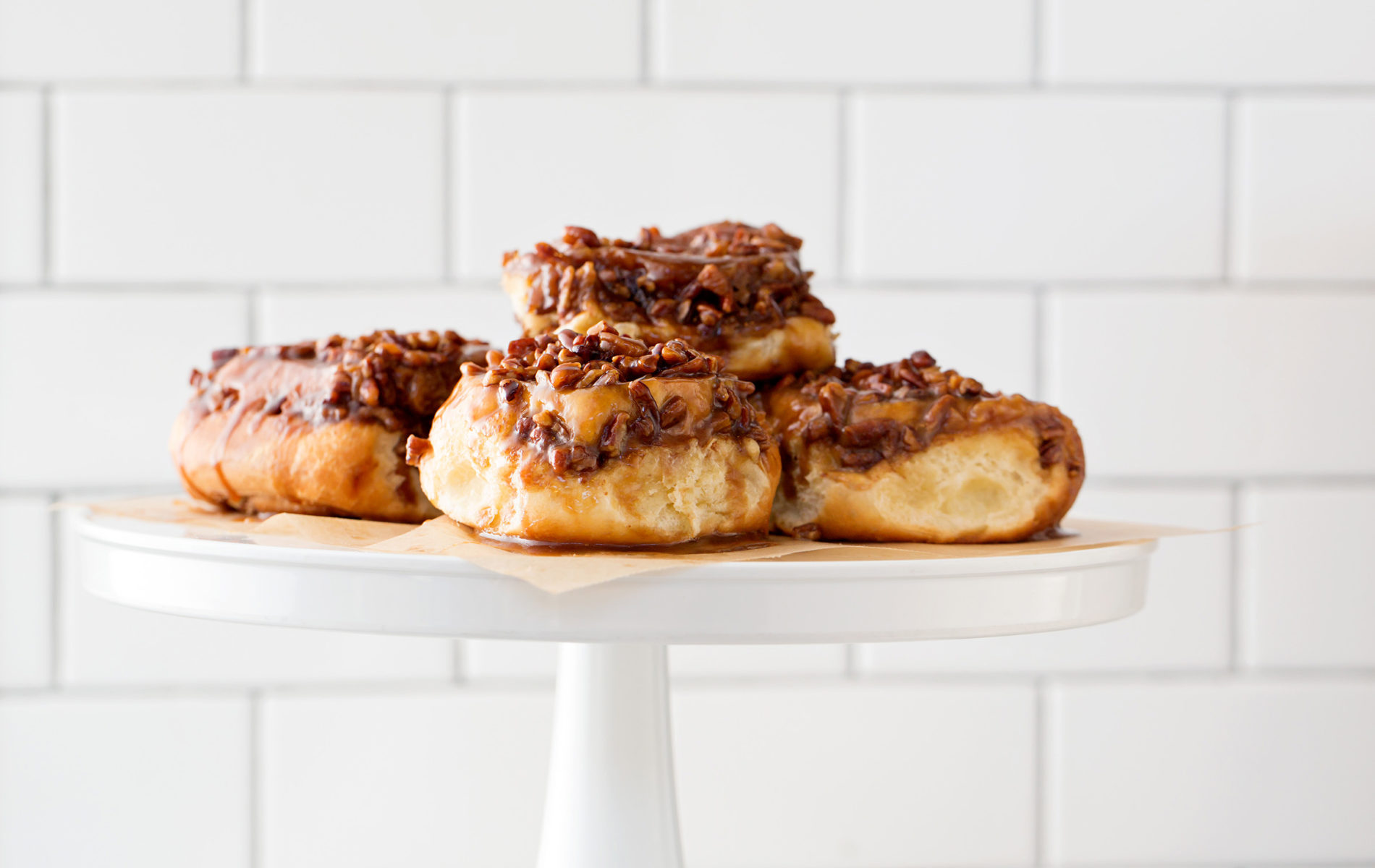a stack of four pecan sticky buns on a white pedestal and a white tile background