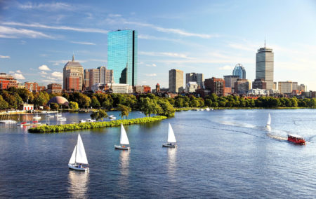 skyline view of boston with a tall blue glass building with river and green park and 3 sail boats