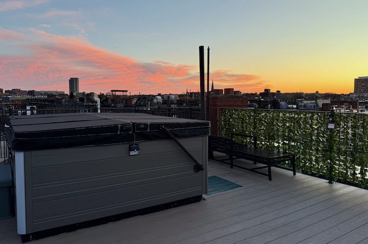 Rooftop deck at sunset with a closed hot tub, a bench, and a green lattice privacy screen against a city skyline.