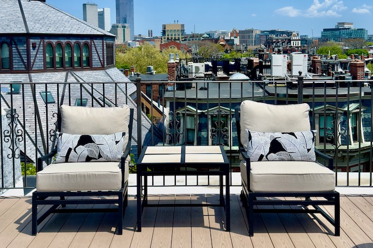 Two cushioned outdoor lounge chairs with a small glass-topped table sit on a wooden balcony overlooking a city skyline.