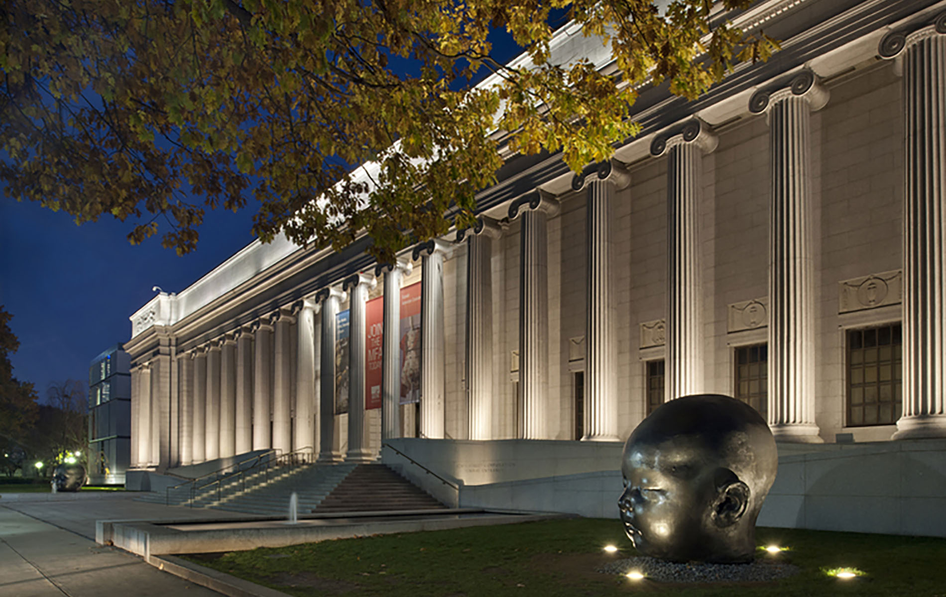 Museum with white columns at night with tree and gaint black state of a head