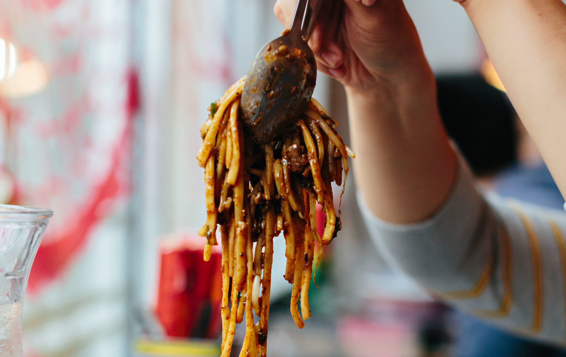 two hands lifting serving spoons full of golden noodles in a sticky brown sauce