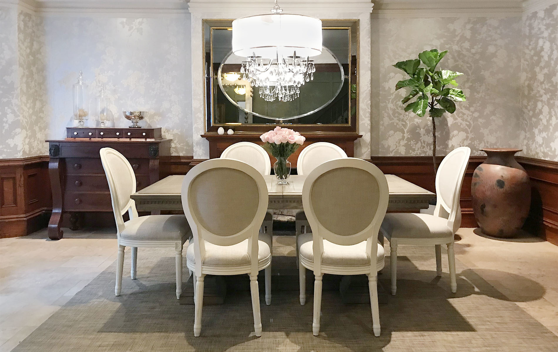 Historic dining room with dark wood wainscot, silver wallpaper with birds and branches, Grey table with six white chairs, pink roses in vase on table.