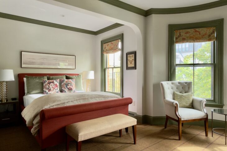 Stylish hotel bedroom with red upholstered bed, green accents, and a tufted white armchair by a window.