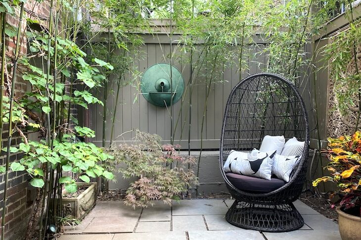 View of bluestone garden, light bronze wooden walls, bamboo, copper water fountain on wall and brown woven basket chair.