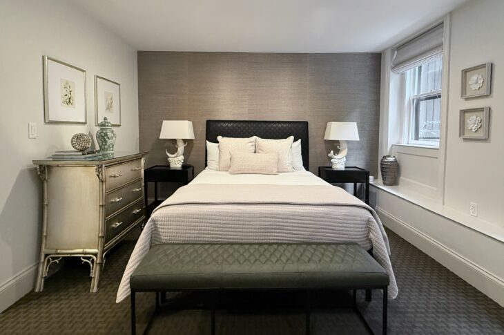 Bedroom with white walls, silver grass cloth wallpaper behind a black headboard. Dark green carpet and window onto garden.