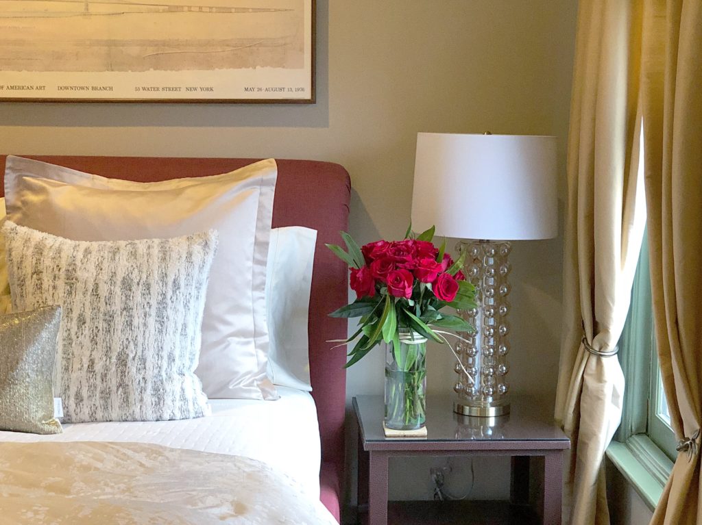 bed with two white and gold pillows with red head board and table with dozen red roses