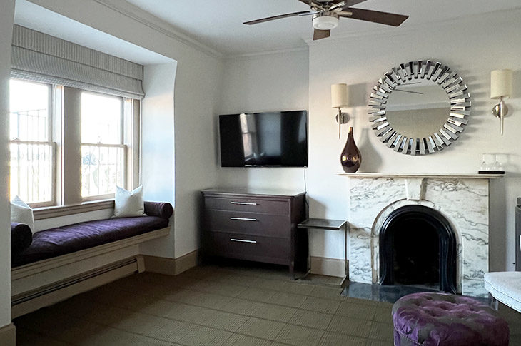 bedroom sitting area, marble fireplace, purple ottoman, brown dresser and padded window seat.