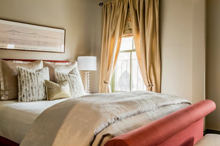 Warm elegant beige room with luxurious bedding, coral colored head and footboards, and window with gold curtains