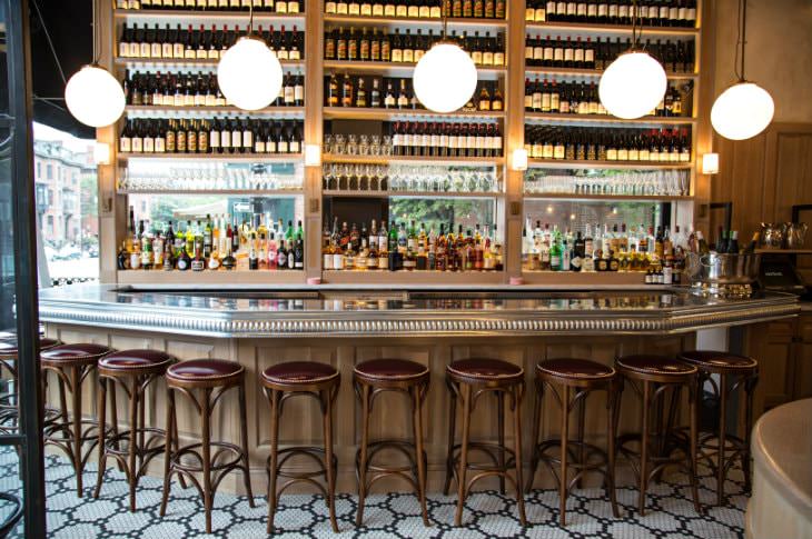 Elegant gold and lightly stained wood bar with round red stools and shelves of wine and liquor bottles and glasses