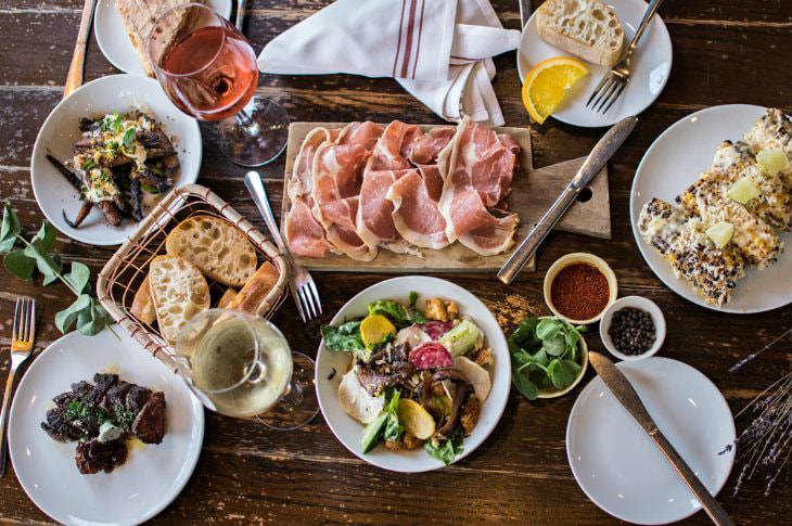 White plates of food, cutting board with meat, and two glasses of rose and white wine on rustic wooden table