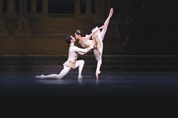 Male ballet dancer on one knee holding a female ballet dancer in Arabesque, both dressed in white and gold