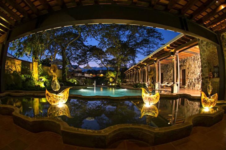 Elegant hotel courtyard swimming pool at dusk surrounded by verandas with stone walls, and tropical plants