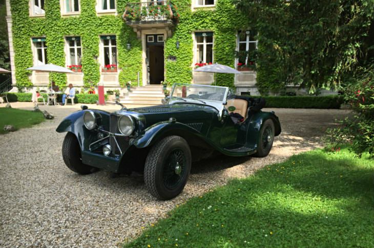 Deep green antique European car on gravel drive with vine-covered hotel in the background