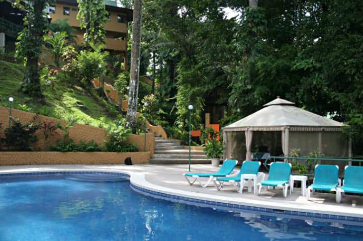 Clear blue swimming pool with turquoise chaise lounge chairs surrounded by lush greenery and a hilltop hotel