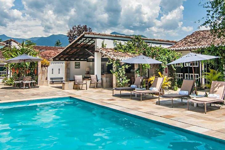 Turquoise swimming pool surrounded by beige tile decking, chaise lounge chairs with umbrellas and buildings with Spanish tile roofs