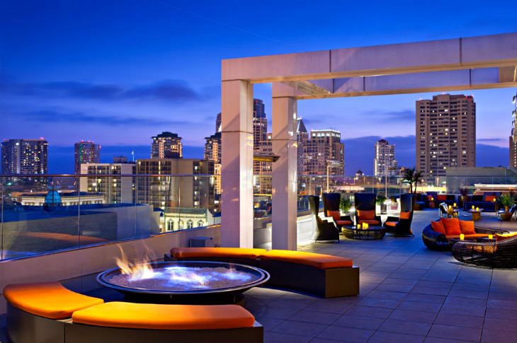 Roof top bar with modern, organically shaped seating and fire pits overlooking a city skyline at dusk