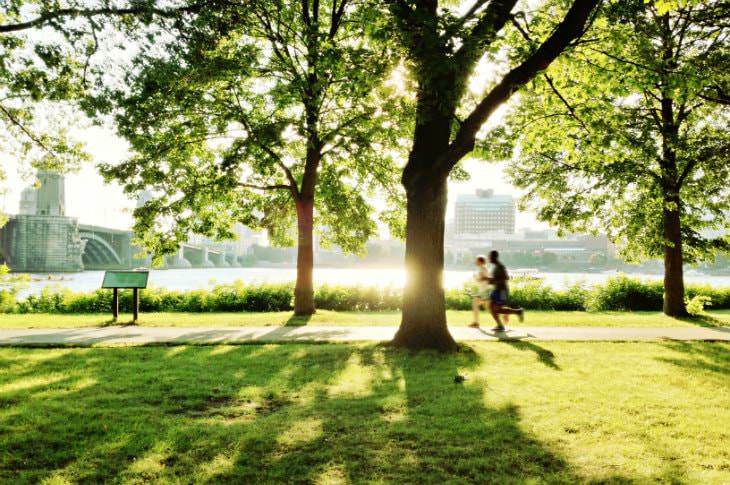 Two people jogging on a path surrounded by green grass and trees with Boston harbor and bridge in the background