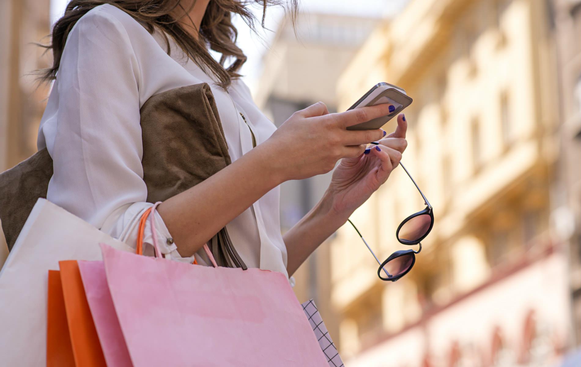 Woman with white, pink and orange shopping bags on her arm, holding her sunglasses, looking at her cell phone