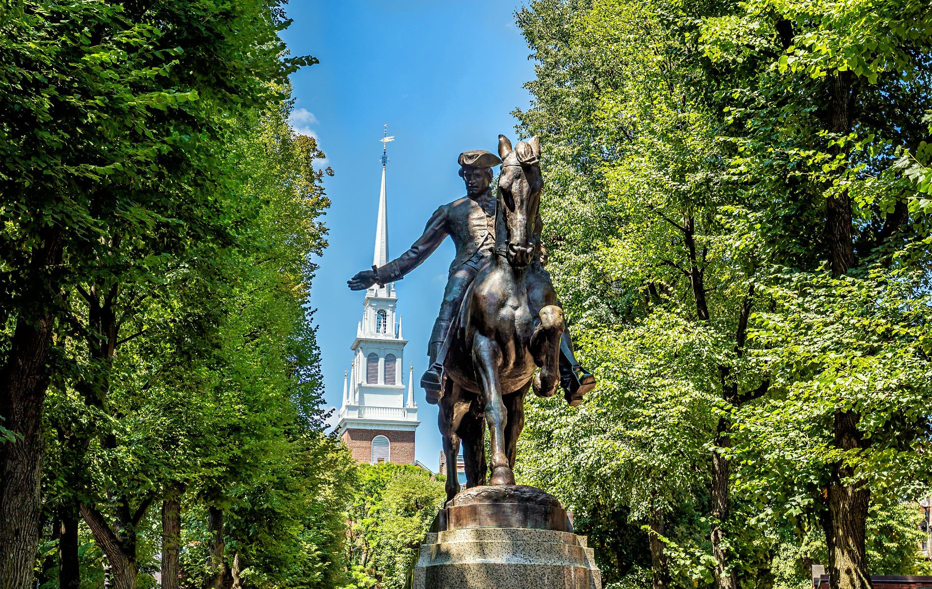 Statue of Paul Revere surrounding by lush green trees with blue skies and a white steeple in the background