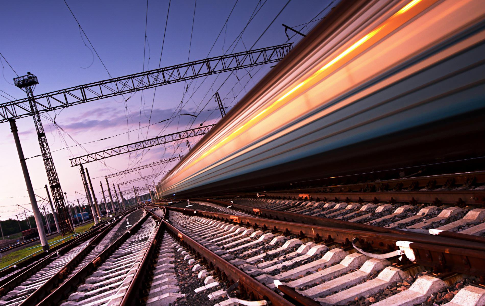 Ground level view of extensive rail system with blurred train racing by amidst purple and pink skies