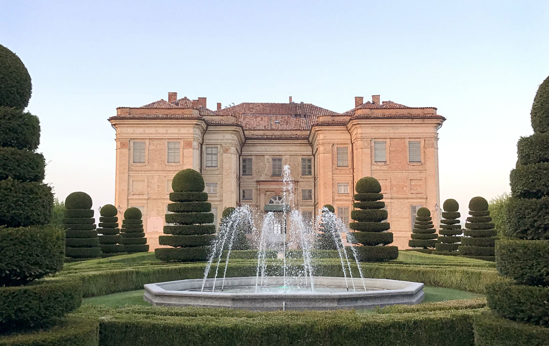 Stone water fountain in formal garden of sculptured shrubs in front of large Italian style building with red tile roof