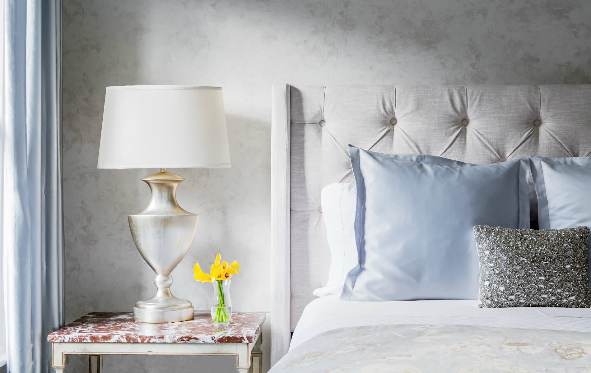 Bed with beige tufted headboard, white sheets and blue pillows, white nightstand with white-washed gold lamp and yellow flowers