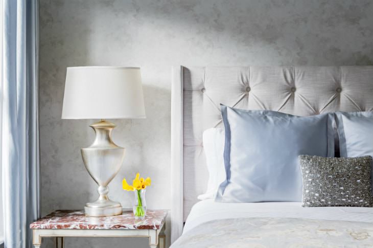 Bed with beige tufted headboard, white sheets and blue pillows, white nightstand with white-washed gold lamp and yellow flowers