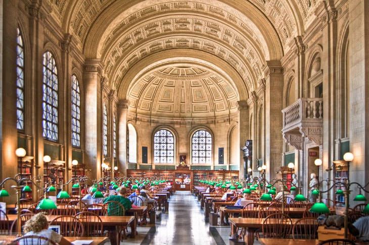 Interior view of library with tall, elaborately carved barrel ceiling, arched windows, and wood tables and chairs