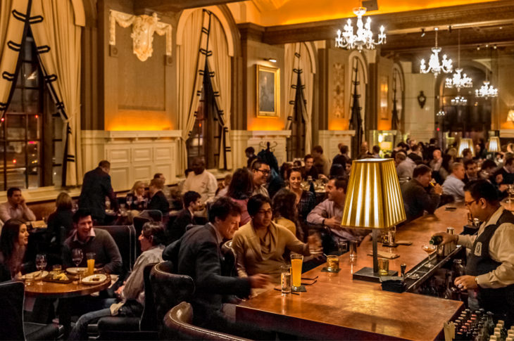 Darkly lit dining room with bar, chandeliers, white heavy drapes and dark upholstered club chairs.