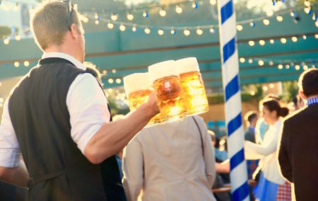 man in black vest carrying 3 large glass beer mugs in party next to a white and blue stripe poll