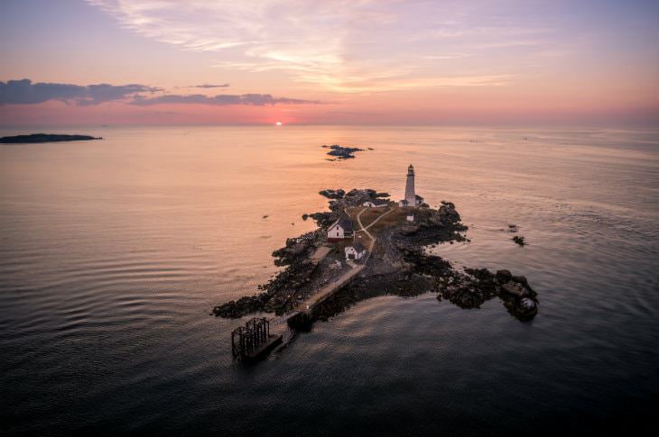 Small island in Boston's harbor with white lighthouse and small buildings amidst purple and pink skies