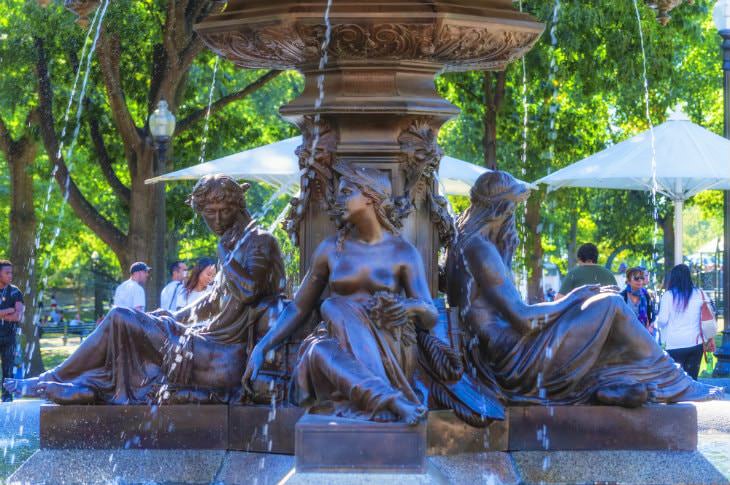 Close-up view of bronze fountain detailing four women around the base with green trees in the background